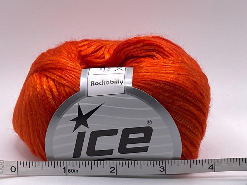 Puede incluir: Una madeja de hilo naranja brillante con una etiqueta "Rockabilly" y la marca "ICE". Una cinta m&eacute;trica debajo muestra una longitud de aproximadamente 10 cm.