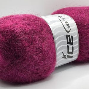 May include: A close-up of two balls of vibrant magenta yarn. The yarn is fluffy and soft, with a label wrapped around the center. The label is white with the brand name "ICE yarns" and a star logo. Ideal for knitting and crochet projects.