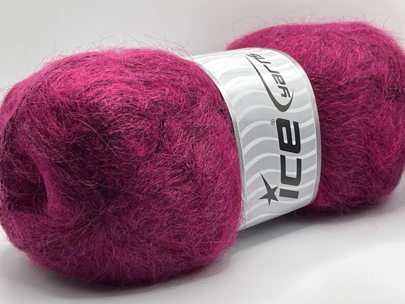 May include: A close-up of two balls of vibrant magenta yarn. The yarn is fluffy and soft, with a label wrapped around the center. The label is white with the brand name "ICE yarns" and a star logo. Ideal for knitting and crochet projects.