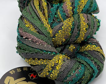 Könnte beinhalten: Ein Knäuel Noro-Garn in Grün-, Grau- und Gelbtönen. Das Garn ist ein strukturiertes Bandgarn mit einer Mischung aus Farben. Ein schwarzes Etikett mit dem Noro-Logo und dem Text "the world of nature Made in Japan" ist angebracht.