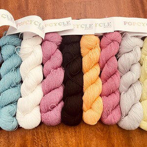 May include: A collection of colorful yarn skeins in various shades, including blue, teal, white, pink, black, orange, mauve, gray, and yellow. Each skein is labeled with the text "POPCYCLE". The yarn is arranged on a wooden surface.