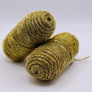 May include: Two golden-yellow yarn skeins, tightly wound and glistening. The yarn has a metallic sheen, suggesting a decorative or craft use. The skeins are positioned on a plain white background, highlighting their texture and color.