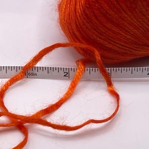 Puede incluir: Una madeja de hilo naranja brillante con un hilo que se extiende hacia la izquierda. Una cinta m&eacute;trica se coloca junto al hilo, mostrando una longitud de aproximadamente 5 cm. El hilo es de un color s&oacute;lido y vibrante.