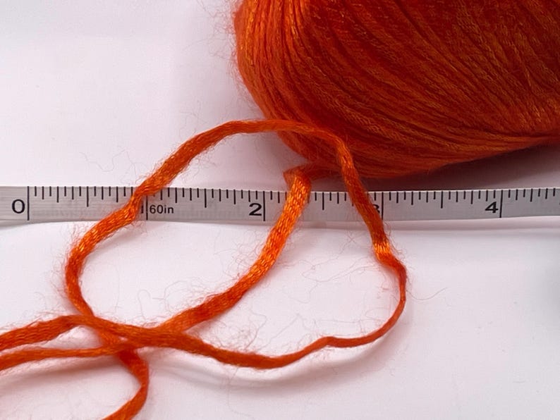 Puede incluir: Una madeja de hilo naranja brillante con un hilo que se extiende hacia la izquierda. Una cinta m&eacute;trica se coloca junto al hilo, mostrando una longitud de aproximadamente 5 cm. El hilo es de un color s&oacute;lido y vibrante.