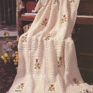 May include: Cream-coloured, crocheted blanket with a textured, bobble stitch design. The blanket features embroidered floral motifs in pink and green. The bottom edge has a fringe. The number "2690-511" is printed on the blanket.