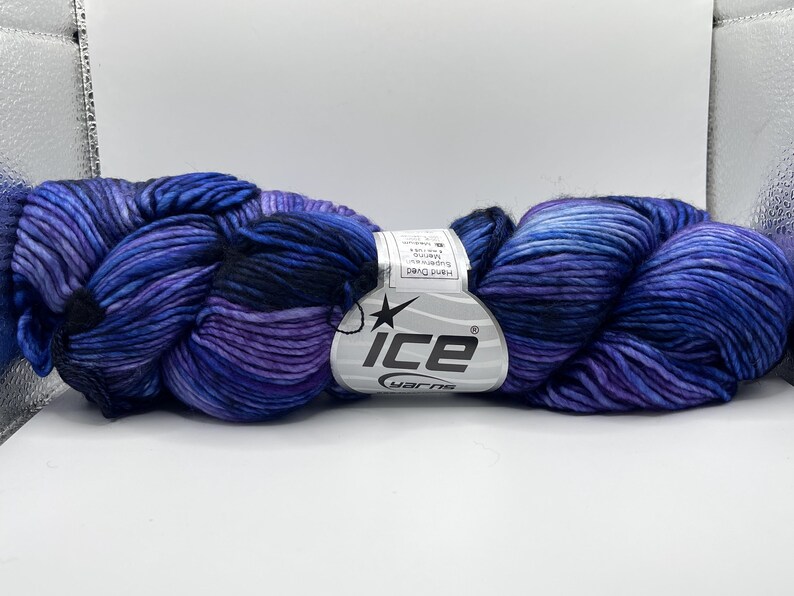 Op de afbeelding: Een streng handgeverfd garen in tinten blauw, paars en zwart. Het garen is gelabeld met "ICE yarns" en lijkt een zacht, getextureerd materiaal te zijn. De kleurveranderingen cre&euml;ren een visueel aantrekkelijk effect.