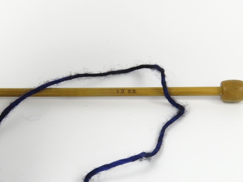 Op de afbeelding: Een houten breinaald met donkerblauw en zwart garen. De naald is gemarkeerd met "5,0 mm". Het garen is over de naald gelust, wat wijst op een breiproject.