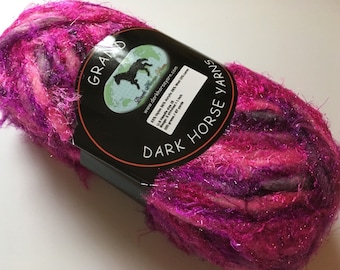 Può includere: Un gomitolo di filato rosa, viola e bianco con fili di glitter argentati. Il filato è etichettato "Dark Horse Yarns" e "Grand".