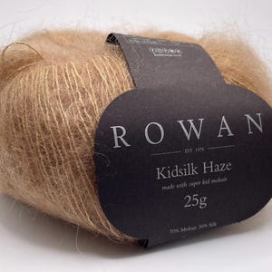 以下が含まれることがあります： ライトブラウン色のRowan Kidsilk Hazeの毛糸玉。ラベルには「ROWAN」と「Kidsilk Haze」と25gの重量が記載されています。この糸は70％モヘアと30％シルクで作られています。
