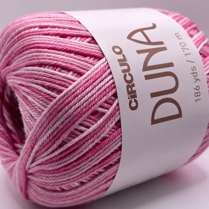 May include: A ball of pink and white variegated yarn with a label that reads "CIRCULO DUNA". The label also indicates the yarn is 186 yds / 170 m. The yarn is tightly wound and appears soft, ideal for knitting or crochet projects.