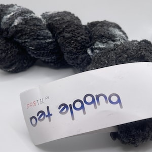 May include: A skein of dark gray yarn with white highlights. The yarn has a textured, bulky appearance. A white label with the words "bubble tea" in blue and the brand "by HIKOO" is in the foreground.