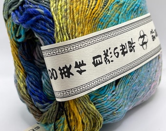 May include: A skein of variegated yarn featuring a blend of yellow, blue, green, and teal fibers. A paper band wraps around the center with Japanese text and a logo. A rectangular tag is attached, displaying the text NORO, The World of nature.