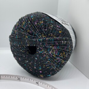 May include: A ball of black yarn with multicoloured metallic threads. The yarn is wound around a white label with black text.