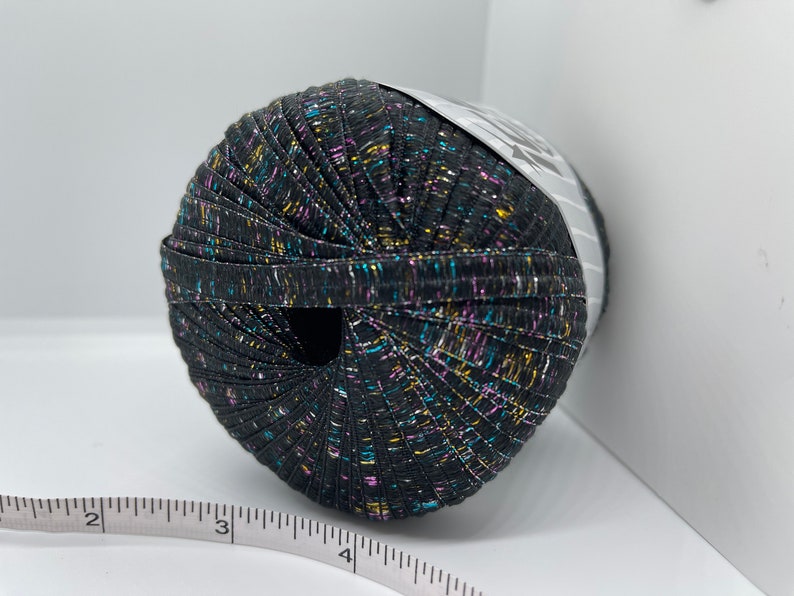 May include: A ball of black yarn with multicoloured metallic threads. The yarn is wound around a white label with black text.