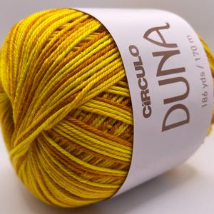 May include: A ball of yellow and orange variegated yarn with a white label wrapped around it. The label reads "CIRCULO DUNA" and "186 yds / 170 m". The yarn is tightly wound and appears soft, suitable for knitting or crochet projects.