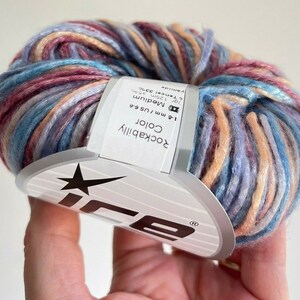 May include: A ball of variegated yarn featuring a blend of blue, pink, and peach tones. A white paper label is wrapped around the center, displaying the text Rockabilly Color, 4-6 mm / US 6-8, and fiber content details including 33% Tencel.