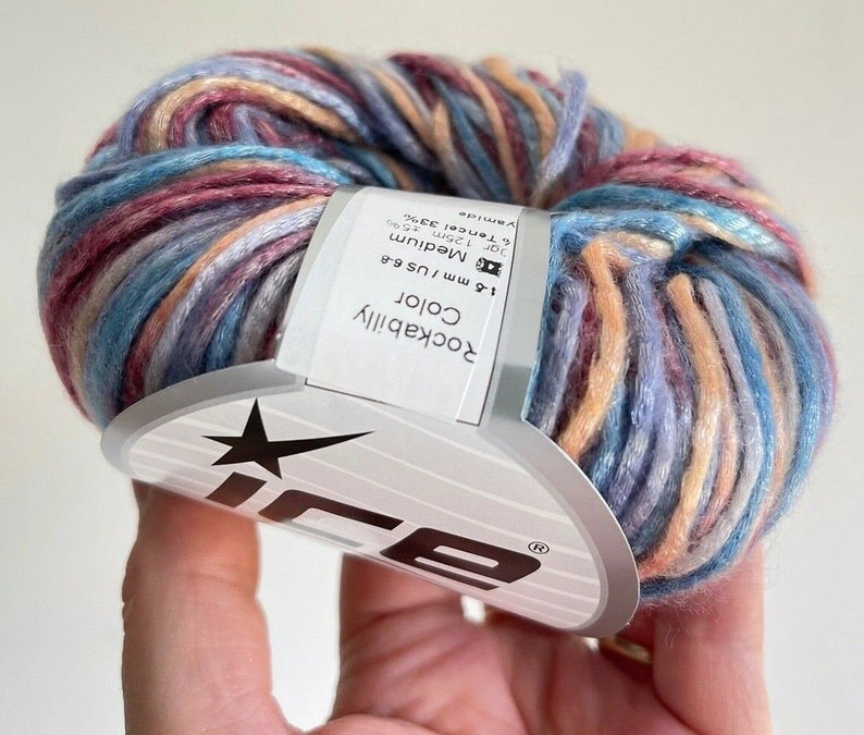May include: A ball of variegated yarn featuring a blend of blue, pink, and peach tones. A white paper label is wrapped around the center, displaying the text Rockabilly Color, 4-6 mm / US 6-8, and fiber content details including 33% Tencel.