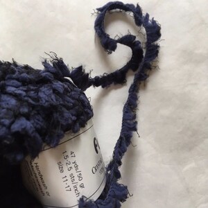 May include: A skein of navy blue yarn with a fringe trim. The yarn label reads "Crystal 1200g 47 yds/50 gr 1.5-2.5 sts/inch size 11-17 Handwash or Machinewash entire cycle".
