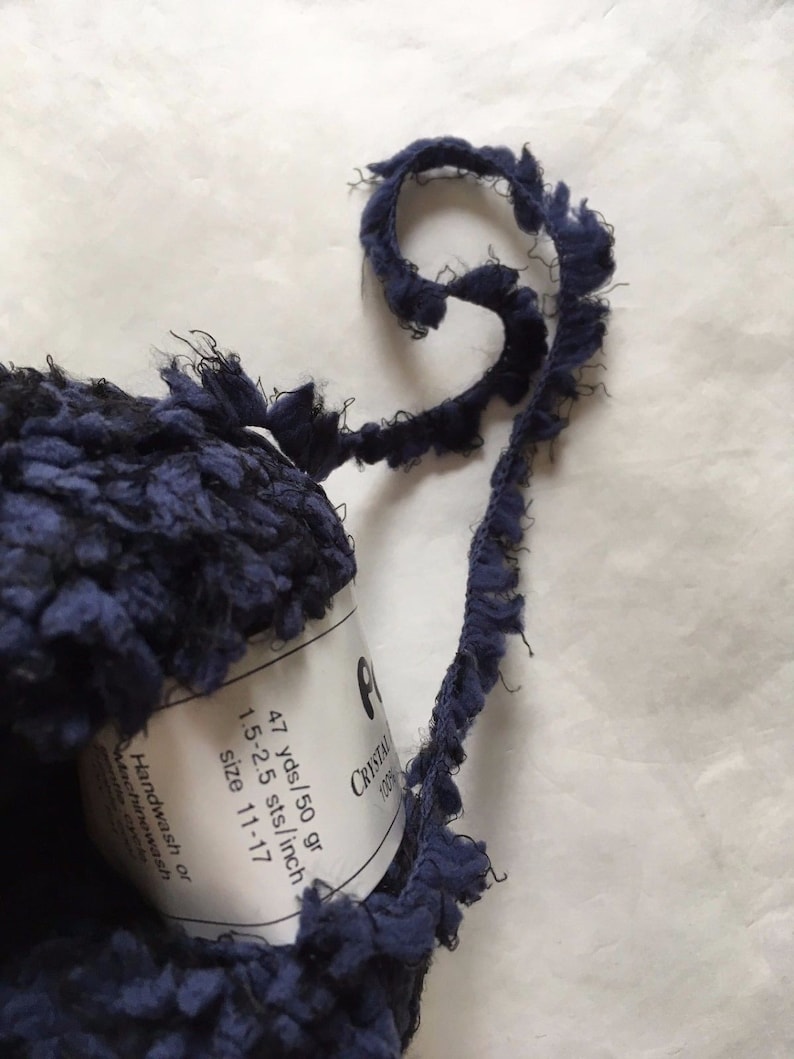 May include: A skein of navy blue yarn with a fringe trim. The yarn label reads "Crystal 1200g 47 yds/50 gr 1.5-2.5 sts/inch size 11-17 Handwash or Machinewash entire cycle".
