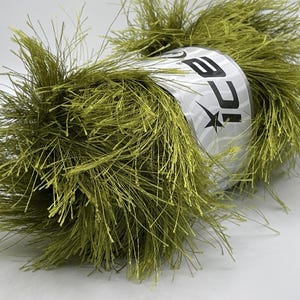 May include: A skein of olive green yarn with a fluffy, textured appearance. The yarn is wrapped around a silver label with the word "ICE" and a star logo. Ideal for knitting and crochet projects.