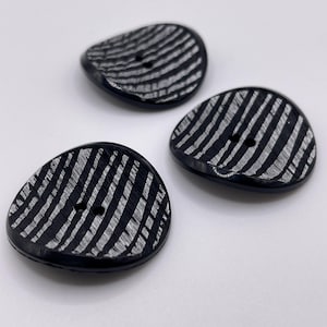 May include: Three black, oval-shaped buttons with a textured, white, striped pattern. Each button has two small holes for sewing. The buttons are arranged on a white surface.