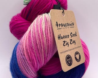 May include: A vibrant skein of hand-painted yarn in shades of pink, blue, and green. The yarn is labeled "Huasco Sock Zig Zag" and is a superwash wool blend. The colors transition smoothly, creating a visually appealing gradient.