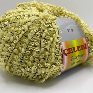 Könnte beinhalten: Ein Knäuel gelbes Garn mit einem strukturierten, geschlungenen Design und schwarzen Fadenakzenten. Das Etikett lautet "CELEBI Purpur" und gibt ein Gewicht von 50 Gramm an. Ideal für Strick- und Häkelprojekte.
