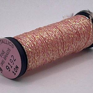 May include: A spool of Kreinik Metallics Fine (#8) Braid thread in a shimmering pink and gold colorway. The spool label displays the text Kreinik Metallics, Fine (#8) Braid, 9192, and 10M. The thread is wound around a black plastic cylinder.