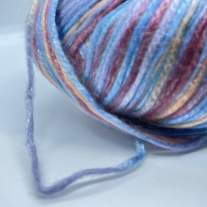 May include: A ball of variegated yarn featuring a blend of light blue, lavender, soft pink, and cream colors. The yarn has a slightly fuzzy, soft texture with a subtle sheen, and a single strand extends from the main ball, showing the twisted fiber.