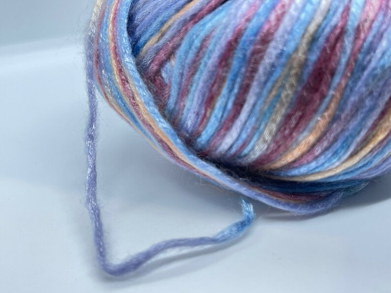 May include: A ball of variegated yarn featuring a blend of light blue, lavender, soft pink, and cream colors. The yarn has a slightly fuzzy, soft texture with a subtle sheen, and a single strand extends from the main ball, showing the twisted fiber.