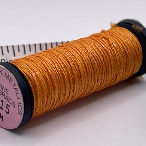 May include: A spool of orange metallic thread labeled Kreinik Metallics Fine (#8) Braids 5515 10M. The spool is positioned in front of a white measuring tape showing a length of approximately 2 inches from the 1-inch mark to the 3-inch mark.