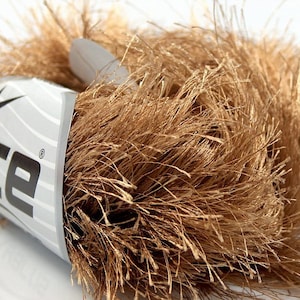 May include: A close-up of a skein of brown yarn with a feathery texture. The yarn is partially wrapped with a label that reads "ICE" in bold, gray letters, with a star graphic. The yarn appears soft and fluffy, ideal for knitting or crochet projects.