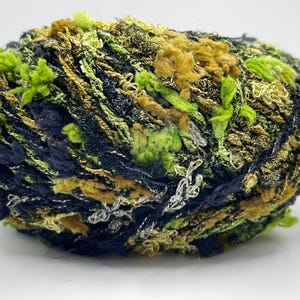 May include: A ball of textured yarn in shades of green, navy, and gold. The yarn has a fluffy, multi-dimensional appearance, with strands of varying textures and colors. Ideal for knitting or crochet projects.