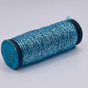May include: A spool of bright metallic blue thread with a textured, braided appearance. The spool end features the text KREINIK METALLICS TAPESTRY (#12) BRAID 014HL 10M. The thread is wound tightly around a black cylindrical core with blue end caps.