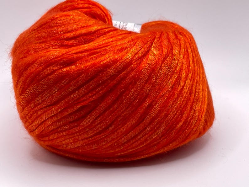 Puede incluir: Una madeja de hilo naranja vibrante, bien enrollada y texturizada. El hilo parece ser una fibra fina y suave, ideal para proyectos de tejido o ganchillo. El color brillante sugiere un producto final audaz y llamativo.