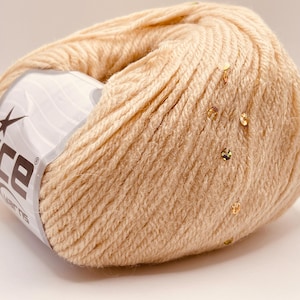May include: A ball of beige yarn with gold sequins scattered across the surface. The yarn is wrapped around a white label with the brand name "ice yarns" and a star logo. Ideal for knitting or crochet projects.