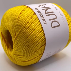 May include: A bright yellow ball of Duna yarn by Circulo. The label indicates 186 yds / 170 m. The yarn is tightly wound, showing a smooth texture. Ideal for knitting and crochet projects.