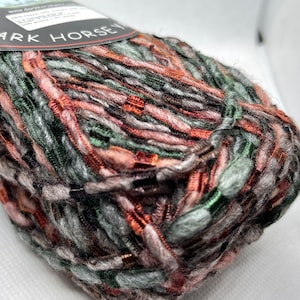 Puede incluir: Una madeja de hilo en una mezcla multicolor de gris, verde, rosa y naranja. El hilo está etiquetado como "Dark Horse" y está hecho de un material suave y esponjoso.