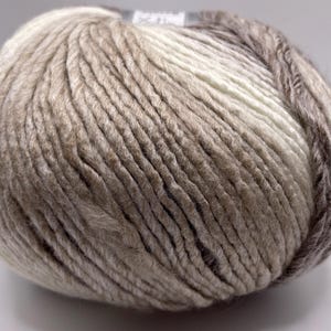 May include: A ball of yarn with a gradient of colors, including white, beige, and brown. The yarn is thick and textured, suitable for knitting or crocheting projects. The ball is tightly wound, showing the individual strands.