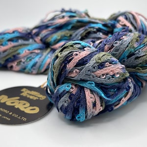 May include: A close-up of a skein of Noro yarn in shades of blue, pink, and green. The yarn has a textured, ribbon-like appearance. A black tag with gold text reads "Noro" and "Made in Japan."