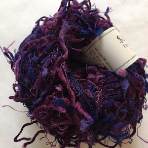 以下が含まれることがあります： 100ヤード/50グラムの紫と青のシルク糸の糸巻き。ラベルには「100 yds/50 grams knit alone on 16 for lacy effect combine off」と書いてあります。編み物用の紫と青の糸です。