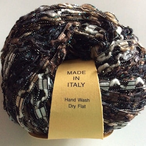 Könnte beinhalten: Ein Knäuel braunes, weißes und goldenes Garn mit silbernen und türkisfarbenen Glitzer. Das Garn ist in Italien hergestellt und sollte von Hand gewaschen und flach getrocknet werden. Das Etikett lautet "Made in Italy" und "Handwäsche Flach trocknen".