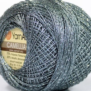 Puede incluir: Una madeja de hilo metálico plateado con una etiqueta que dice "YarnArt Camelia" e incluye la composición: 70% poliéster, 30% metálico. La etiqueta también incluye el texto "YarnArt Camelia" y el peso: 20 g/20/0,700.