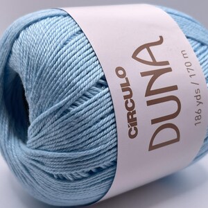May include: A ball of light blue yarn with a white label wrapped around it. The label features the word "DUNA" in large brown letters, along with the brand name "CIRCULO". The label also indicates the yarn is 186 yards / 170 m.