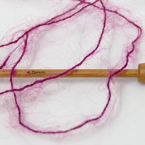 May include: Close-up of a wooden knitting needle with 4.0mm text and a skein of magenta yarn. The yarn is loosely arranged, showing its fluffy texture. The needle is made of light-colored wood.