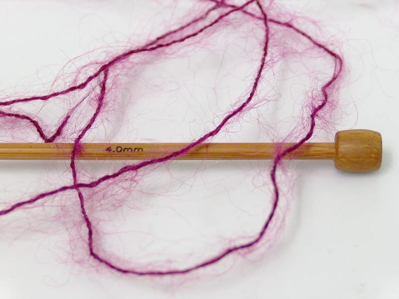 May include: Close-up of a wooden knitting needle with 4.0mm text and a skein of magenta yarn. The yarn is loosely arranged, showing its fluffy texture. The needle is made of light-colored wood.