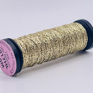 May include: A spool of gold metallic thread featuring a pink end cap with the text KREINIK METALLICS, FINE BRAIDS, (#8) 017HL, and 10M. The thread is wound tightly around the black plastic spool, displaying a shimmering, textured surface.
