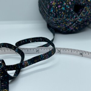 May include: A black ribbon with gold, blue and pink flecks is measured with a white tape measure. The ribbon is 7.6 cm long.