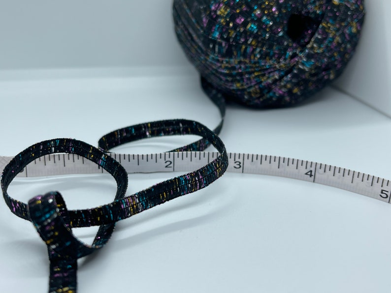 May include: A black ribbon with gold, blue and pink flecks is measured with a white tape measure. The ribbon is 7.6 cm long.