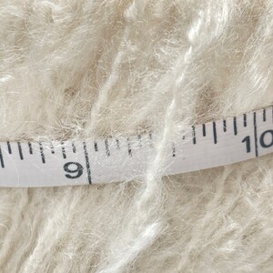May include: A close-up of a white yarn skein with a measuring tape wrapped around it. The tape reads "9" and "10".